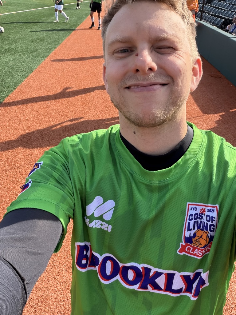 A man on an athletic field, smiling and wearing a soccer jersey that says Brooklyn on the front, with a badge that says 'Cost of Living Classic'