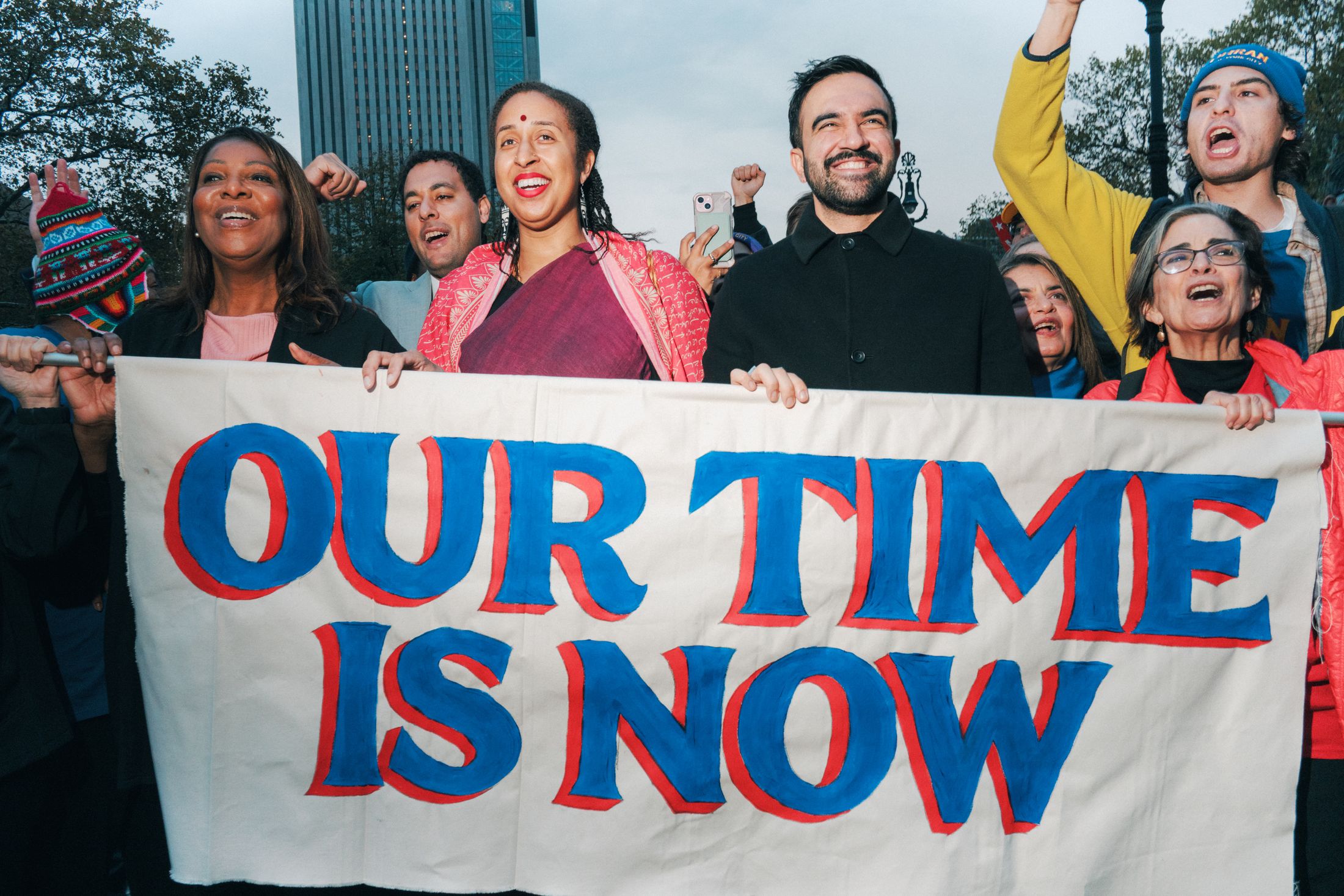 Zohran Mamdani leading a march holding a hand-painted banner that reads 'Our Time is Now' using the Zohran font.